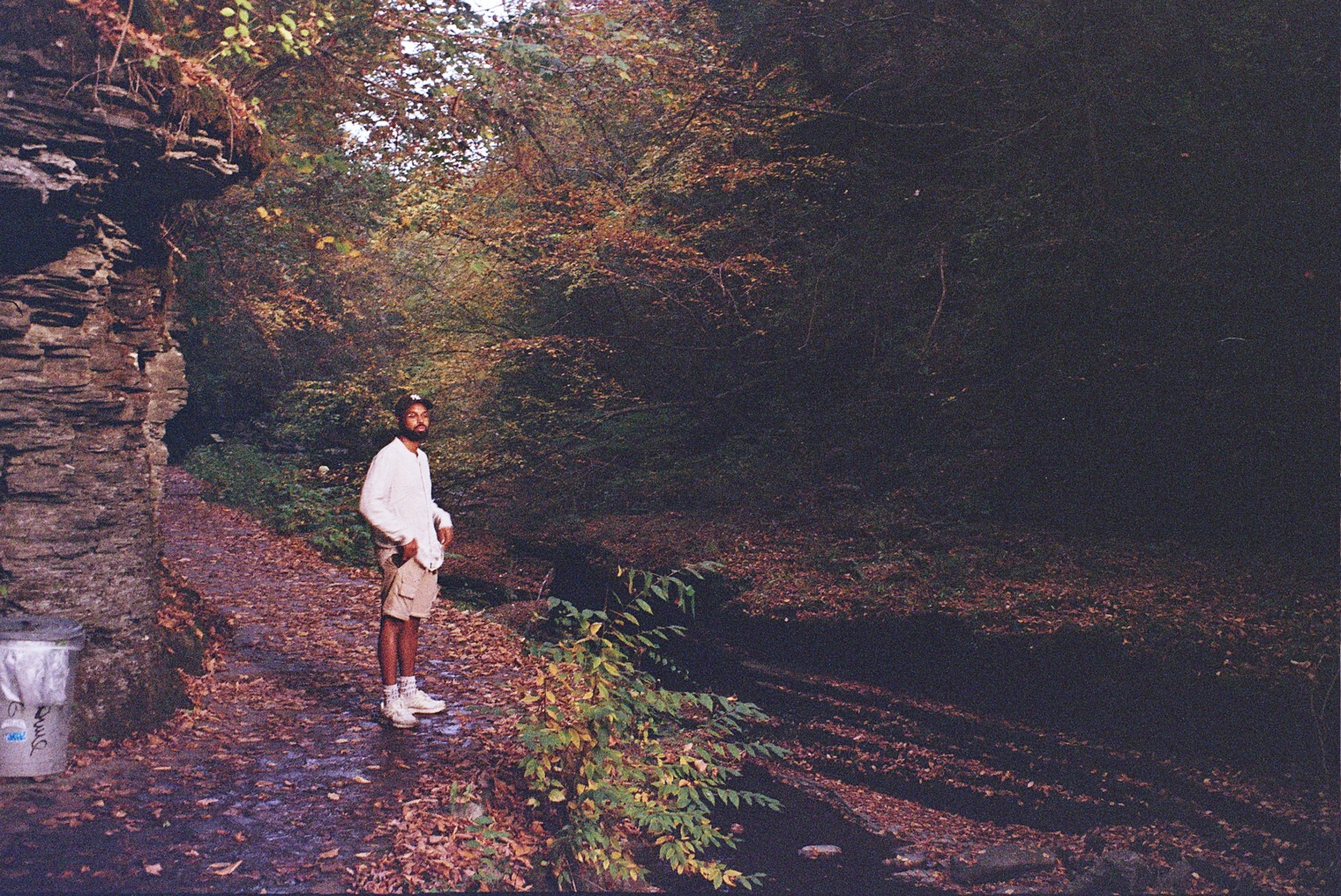 Adé Hakim standing beside a stone wall on a wooded path.