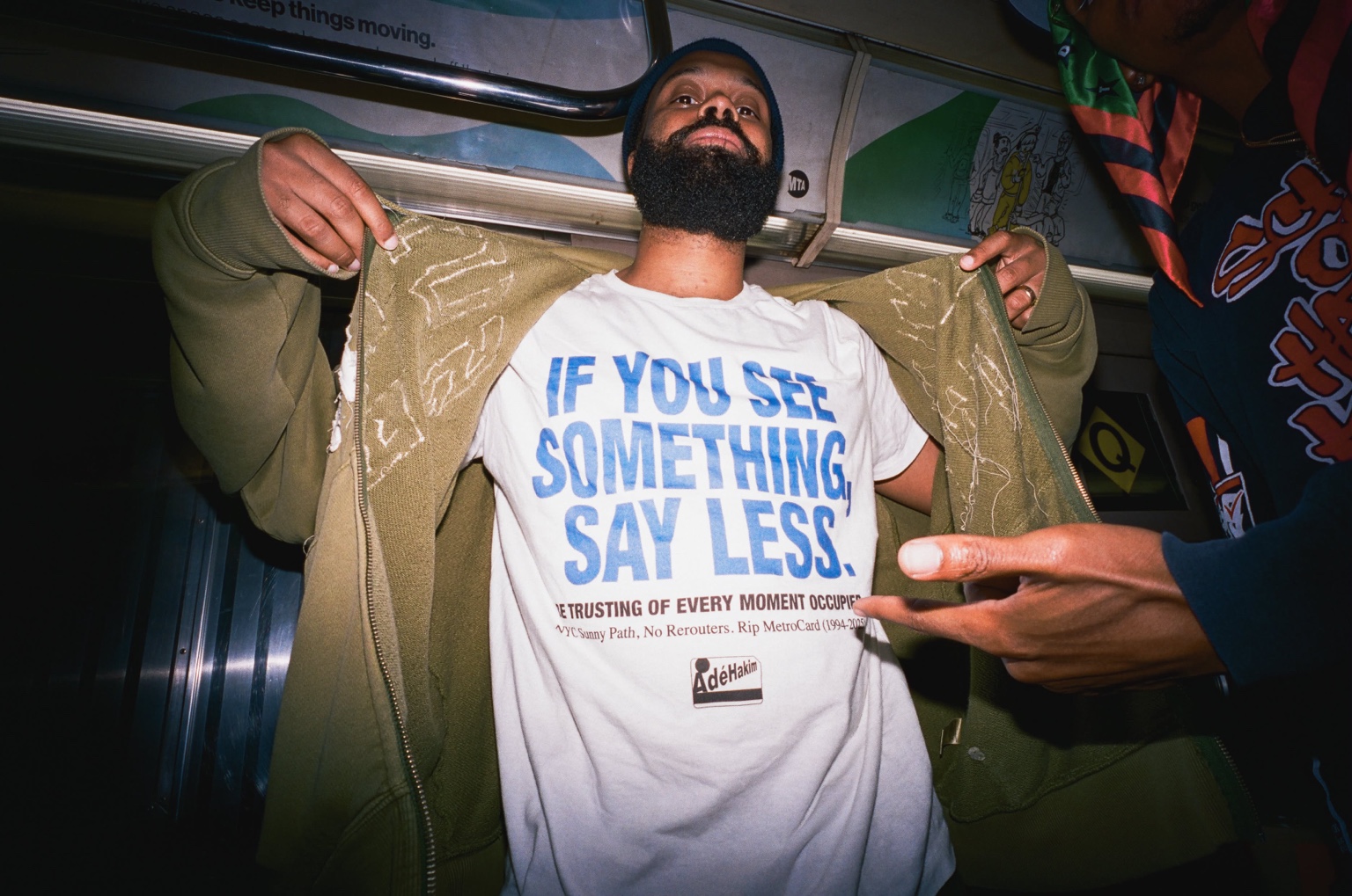 Adé Hakim opens his jacket on the subway to show a shirt reading If you see something, say less.