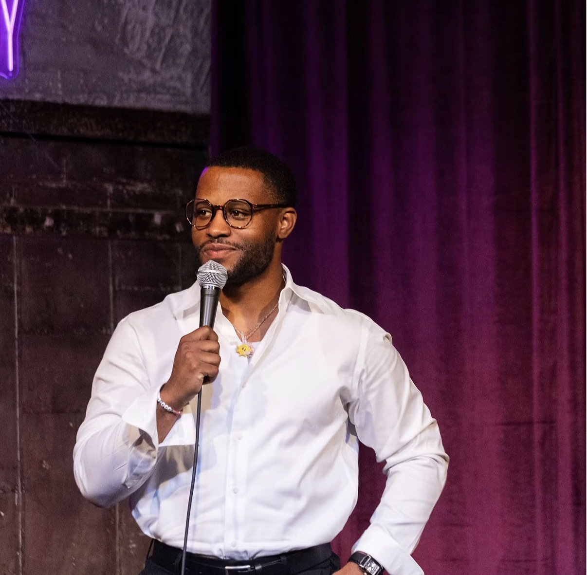 Nassir Saleem performs stand-up with a microphone against a purple-lit curtain