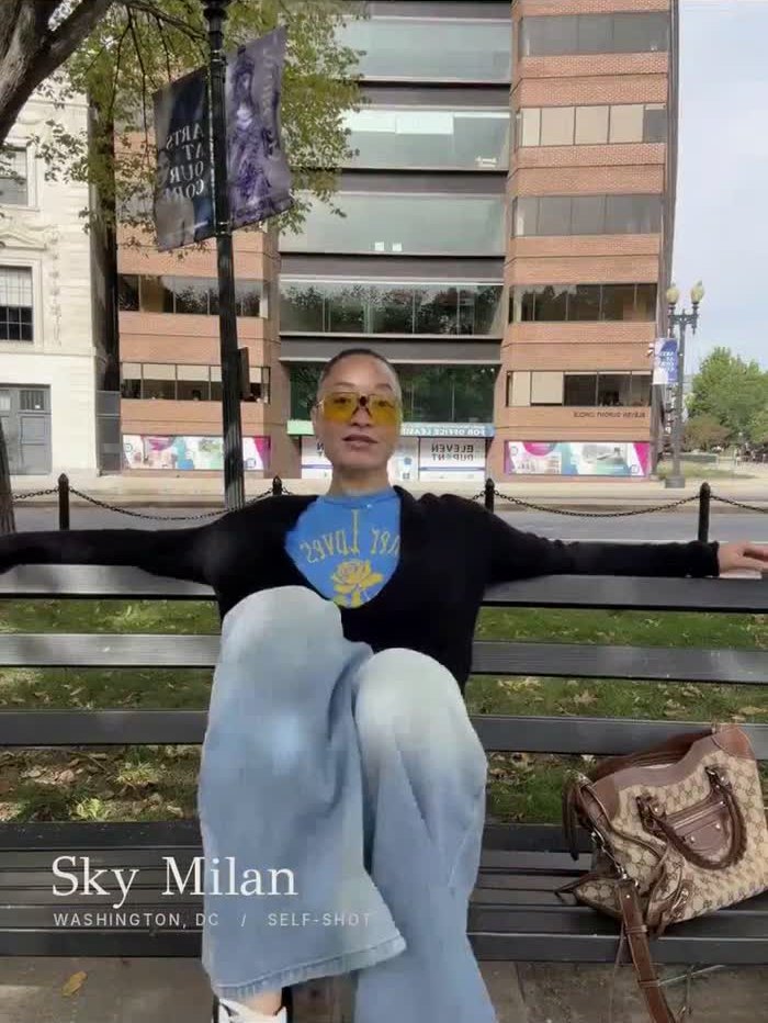 Sky Milan seated on a bench wearing yellow sunglasses and a blue top, in a frame from her SELF-SHOT.
